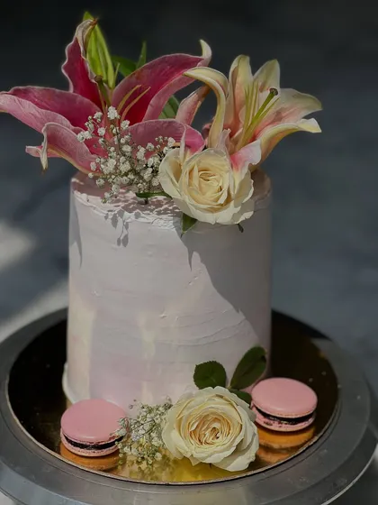 A tall, elegant cake decorated with beautiful pink lilies, white roses, and delicate baby's breath. A couple of macarons at the base add a playful, modern touch to this floral design.
