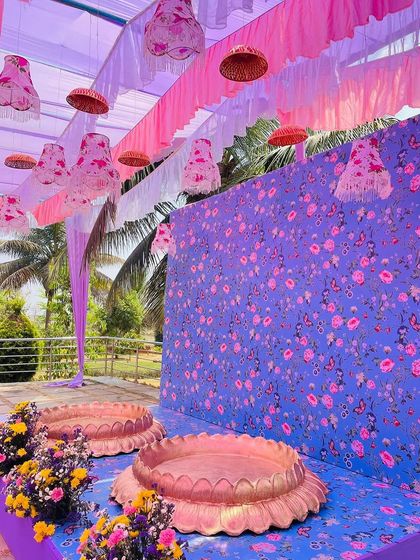 A unique Haldi setup with a purple and pink color scheme. The floral printed backdrop and hanging lampshades create a whimsical and unconventional look.
