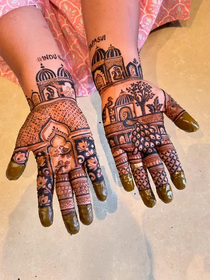This intricate design combines architectural elements with nature. It features palace domes, trees, and a peacock, with one hand showcasing a beautiful floral mandala. The word "Tapasvi" is also included for a religious occasion.
