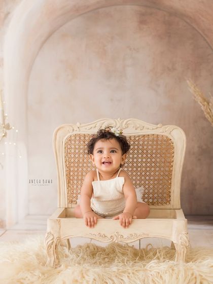 A smile fit for a queen. This adorable baby girl sits happily in an elegant, vintage-style chair, the star of her milestone photoshoot.