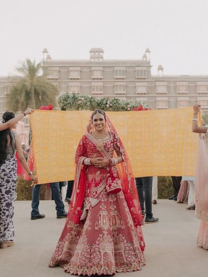 The bride's grand entrance, framed against the majestic facade of the wedding venue, as her bridesmaids hold a traditional cloth.