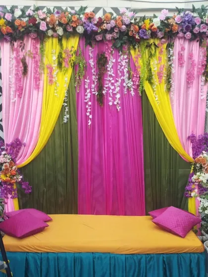 A colorful seating arrangement with a backdrop of pink, yellow, and green drapes. The hanging floral strings add a delicate touch to this vibrant setup for a Mehandi or Haldi.