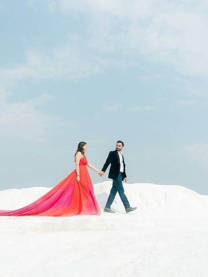 A wide-angle shot from a pre-wedding photoshoot, capturing the couple walking hand-in-hand against a vast, minimalist landscape. The long trail of the gown adds a sense of scale and drama to this romantic portrait.