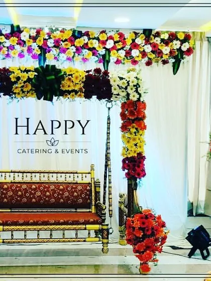 An elegant swing (jhula) setup for a baby shower. The golden swing is decorated with pillars of red and yellow flowers against a simple white curtain backdrop.
