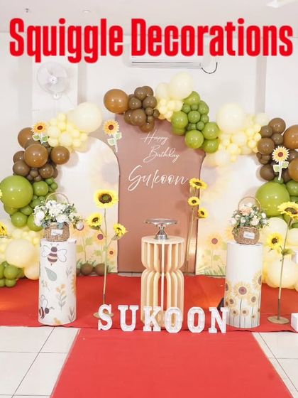 A sunny and cheerful sunflower-themed first birthday party. The decoration features a rustic backdrop, sunflower props, and a balloon arch in shades of green, yellow, and brown.