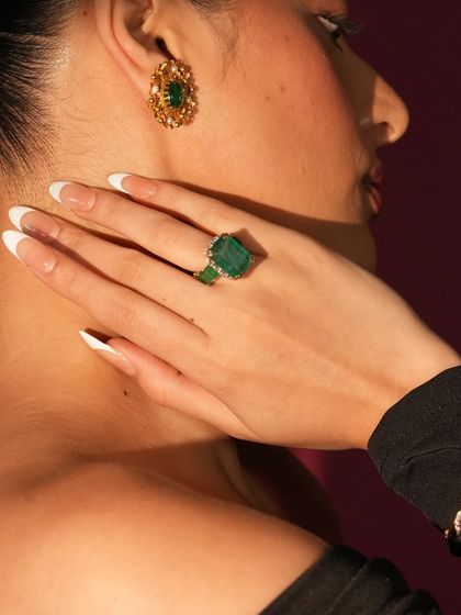 A close-up of a hand adorned with a large emerald cocktail ring and matching gold and emerald earrings.