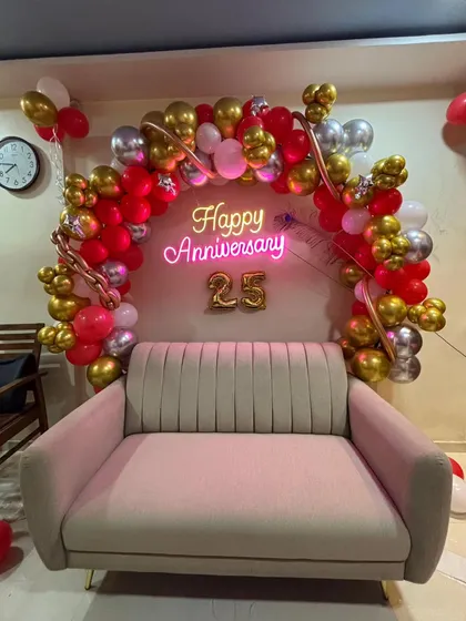A cozy and romantic 25th-anniversary setup for a home. We created a balloon arch in red, gold, and pink over a sofa, with a 'Happy Anniversary' neon sign and '25' foil numbers.
