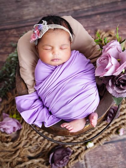 An overhead shot of a newborn wrapped in a purple swaddle, sleeping in a basket surrounded by rustic rope and flowers.