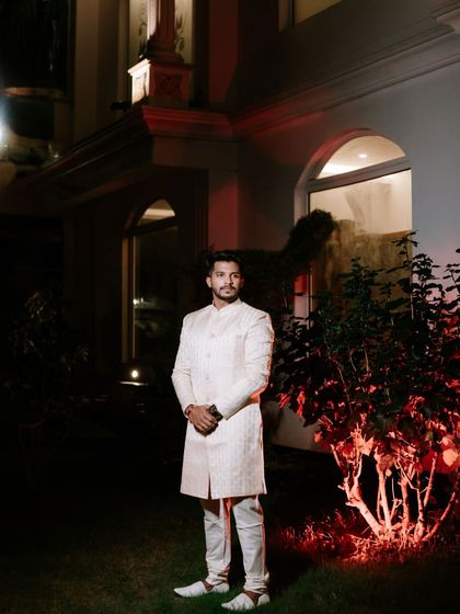 A nighttime portrait of the groom in his elegant sherwani. The warm red lighting from the side adds a touch of drama and highlights his stylish attire.