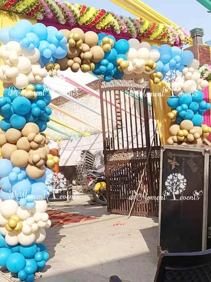 An outdoor balloon arch in blue, beige, and white for the twins' welcome party, set against a backdrop of traditional yellow and pink marigold decorations.