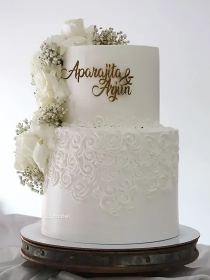 Another view of the all-white two-tier cake, highlighting the personalized gold topper and the beautiful contrast between the smooth top tier and the textured bottom tier.