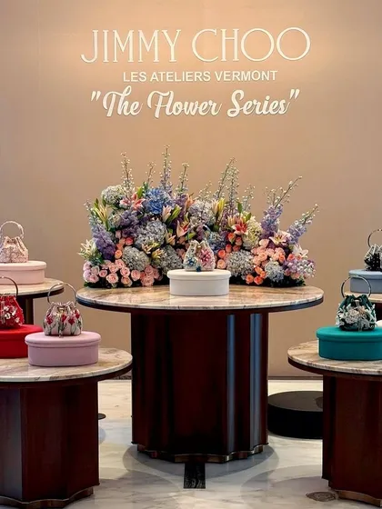 A multi-level product display showcasing Jimmy Choo's new collection. Each handbag is presented on its own table, surrounding a central, grand floral arrangement that unifies the entire presentation.