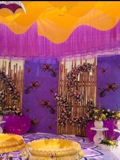 The main seating area for a Haldi ceremony, featuring a unique backdrop with bamboo elements and floral arrangements against a purple fabric. The yellow draped ceiling adds a canopy effect.