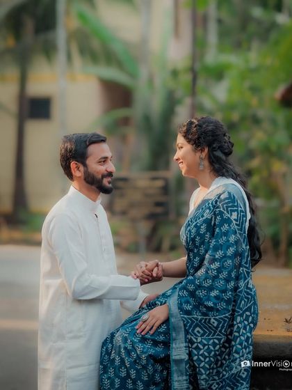 A sweet, interactive moment during a traditional pre-wedding shoot. The groom-to-be stands and holds his partner's hand as she sits, both sharing a happy, loving gaze.