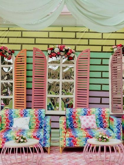 A vibrant and playful photo wall for a carnival-themed Mehendi. The backdrop features a colorful brick pattern with multi-hued window shutters, complemented by rainbow-print sofas for a fun and eclectic look.