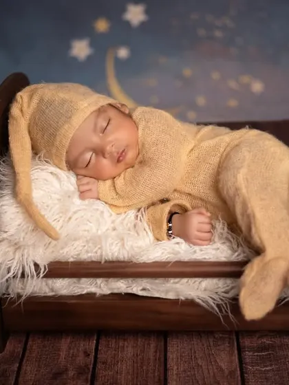 Dreaming of the moon and stars, this newborn sleeps in a miniature bed. The cozy yellow sleeper and starry night backdrop make for a whimsical and sweet scene.