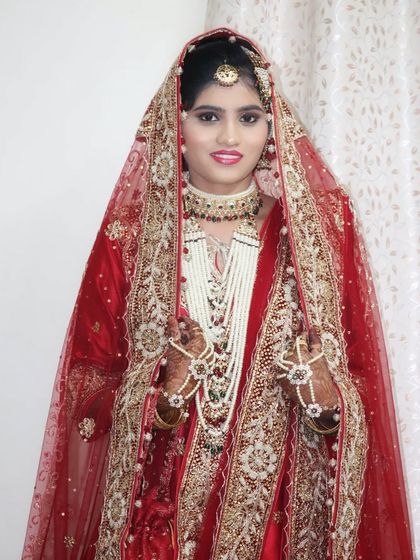 A beautiful Muslim bride in a classic red and gold wedding outfit. The makeup is soft and radiant, with a focus on enhancing her natural beauty, and the look is completed with traditional pearl jewellery.
