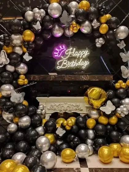 A birthday party with a black, gold, and silver balloon arch. The decoration includes a "Happy Birthday" neon sign with a crown and butterfly accents.