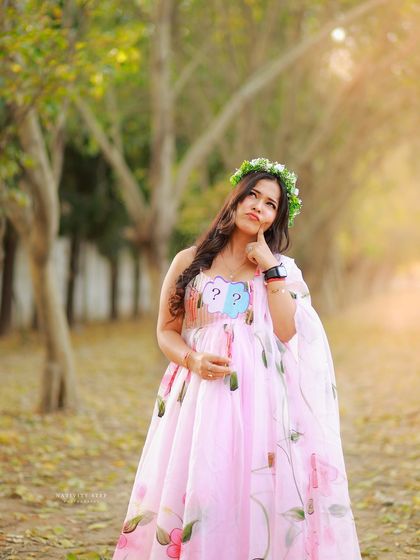A playful outdoor shot with a "boy or girl?" prop. The hand-painted pink gown and floral crown add a whimsical, artistic touch.