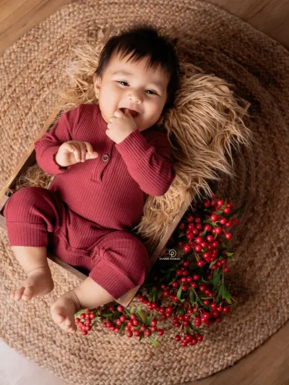 A happy baby in a maroon romper lies in a wooden crate on a jute rug. The simple, rustic props and pops of red berries create a warm and earthy portrait.