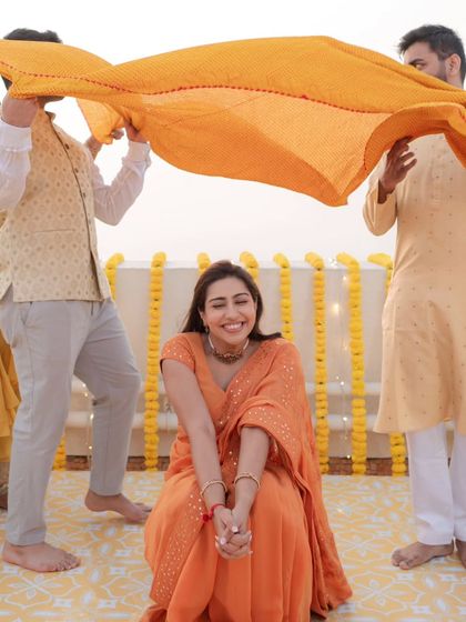 A candid moment of the bride, seated and smiling, as her family playfully holds a yellow dupatta over her head.
