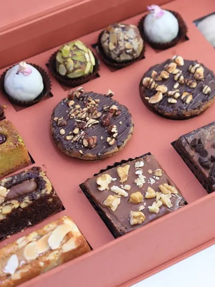 An open box revealing a rich assortment of financier bars, brownies, tarts, and truffles. This is a decadent collection perfect for any Eid dessert table.