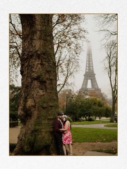 A sweet, cozy portrait of a couple by a large tree in Paris, with the Eiffel Tower visible in the misty background. The image has a vintage, timeless quality.