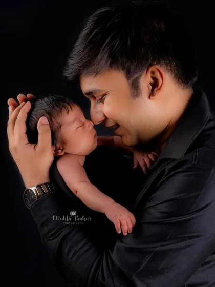 The tender, unbreakable bond between a father and his newborn daughter, captured in a classic, intimate studio portrait against a simple black backdrop.