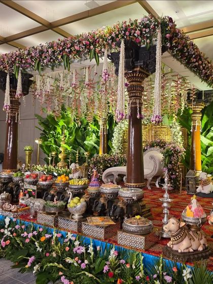 A grand mandapam design featuring traditional carved wooden pillars and a canopy covered in flowers. The backdrop of lush greenery makes the floral colors pop.