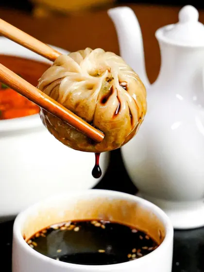 A single dimsum held by chopsticks, dripping with soy sauce. This macro shot captures a perfect, crave-worthy moment, emphasizing the juicy filling and savory sauce.