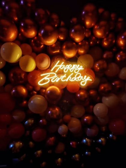 A close-up of the 'Happy Birthday' neon sign nestled within a heart-shaped balloon arrangement. The warm orange glow creates a cozy and inviting atmosphere for the celebration.