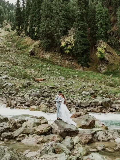 A wide shot capturing the grand scale of the Kashmir landscape as a couple stands by a river. This image highlights the epic, adventurous feel of a destination pre-wedding shoot.