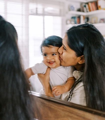 A mother kissing her smiling baby, reflected in a mirror. A creative and intimate shot from a lifestyle session.