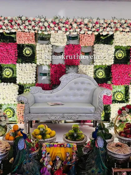 A unique checkerboard backdrop made with panels of pink, white, and green flowers. The silver sofa and a full display of offerings with peacock figures create a modern yet traditional look.