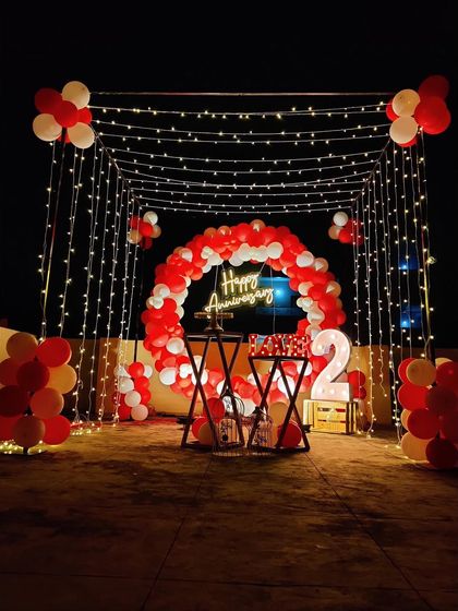 A beautiful terrace setup for a second anniversary. The decoration features a red and white balloon circle, a "Happy Anniversary" neon sign, and a canopy of fairy lights creating a romantic ambiance.