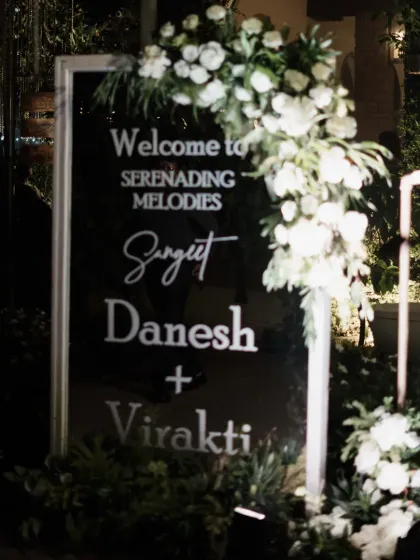 A sleek and modern welcome sign for the "Serenading Melodies" Sangeet. The black acrylic with white lettering, adorned with fresh white flowers, set a sophisticated tone right from the entrance.