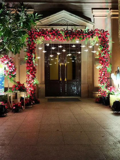 A dramatic night-time entrance featuring a square arch completely covered in red roses. Hanging bird-shaped lights add a whimsical and enchanting touch to the bold floral design.