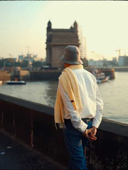 A man in a hat and scarf looks out towards the Gateway of India. This candid back-shot captures a moment of tourism or quiet reflection, with the iconic landmark softly focused in the background.