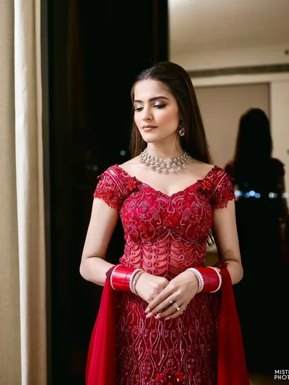 A candid moment by the window. The soft, natural light beautifully highlights the flawless makeup and the intricate details of her red reception gown.
