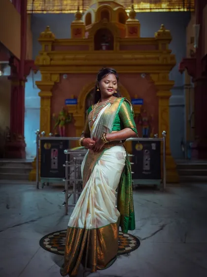 A full-length portrait of a woman in a stunning white and green South Indian saree, standing in the center of a temple hall. The architecture of the temple provides a beautiful, traditional backdrop.