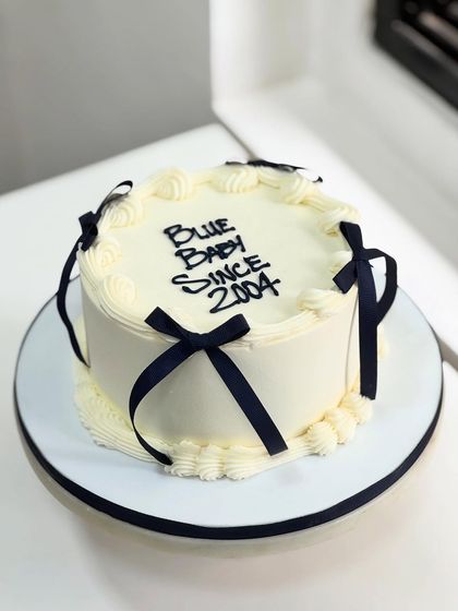 "Blue Baby Since 2004," but make it edible. This Korean-style vintage cake is minimal, bold, and personal, featuring clean white frosting and striking navy blue ribbons.