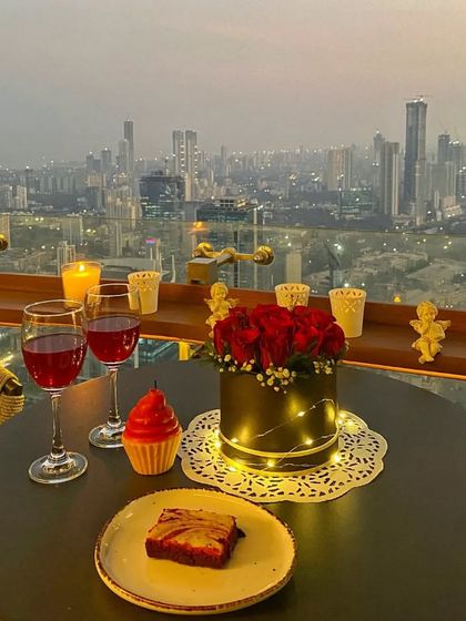 The same terrace setup at dusk, with the city lights beginning to twinkle. The warm glow of the candles and fairy lights on the rose bouquet creates a magical and romantic atmosphere.