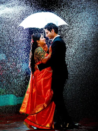 A classic romantic movie moment recreated with artificial rain. The bride's red saree and the groom's suit look stunning in this dramatic pre-wedding shot.