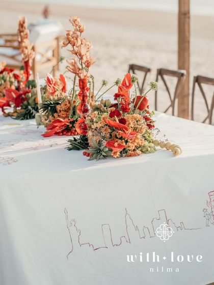 A long table set for a beachside dinner, with a runner of tropical flowers and red glass vases.
