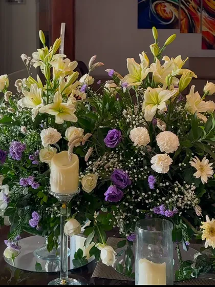 A centerpiece featuring white lilies, purple roses, and baby's breath. The soft glow of the candle adds a touch of warmth to this elegant floral arrangement.