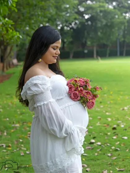 A stunning solo portrait of the mama-to-be in a white off-shoulder dress, holding a bouquet of pink roses. She looks serene and beautiful.