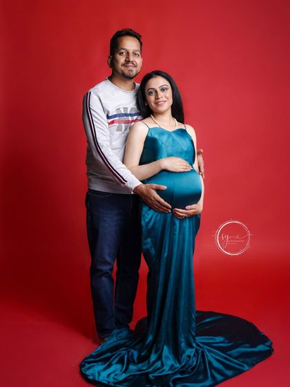 A classic couple's maternity pose. The rich teal gown stands out beautifully against the red background, creating a visually stunning portrait.