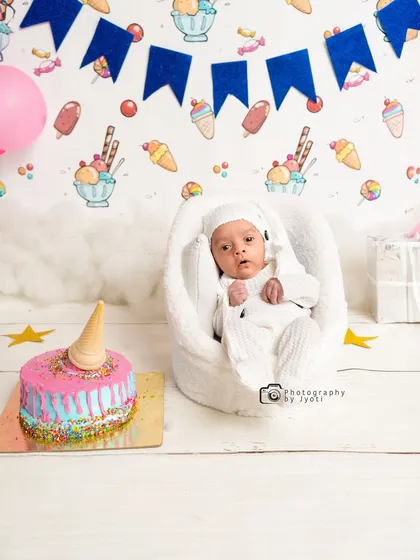 An adorable ice cream-themed milestone shoot. This newborn is surrounded by sweet decorations, including a colorful fake cake and an ice cream-patterned backdrop.