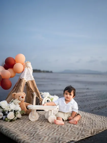 A beautiful solo portrait of a baby boy during his first birthday beach shoot. The boho-inspired setup with a teepee, teddy bear, and balloons creates a whimsical scene.