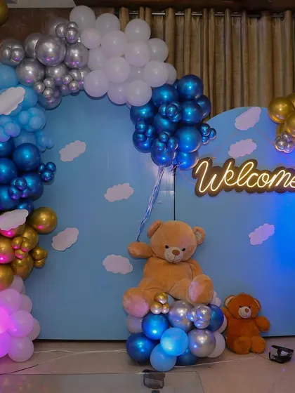 A classic "Welcome Home Baby" decoration designed for an indoor space. This setup includes a large teddy bear, a custom neon "Welcome" sign, and a beautiful balloon arch in shades of blue, silver, and gold against a sky-themed backdrop.